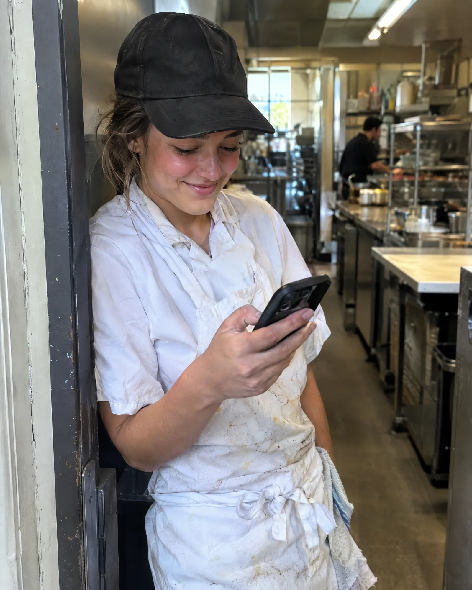 A line cook smiling at a text on her phone in the kitchen doorway
