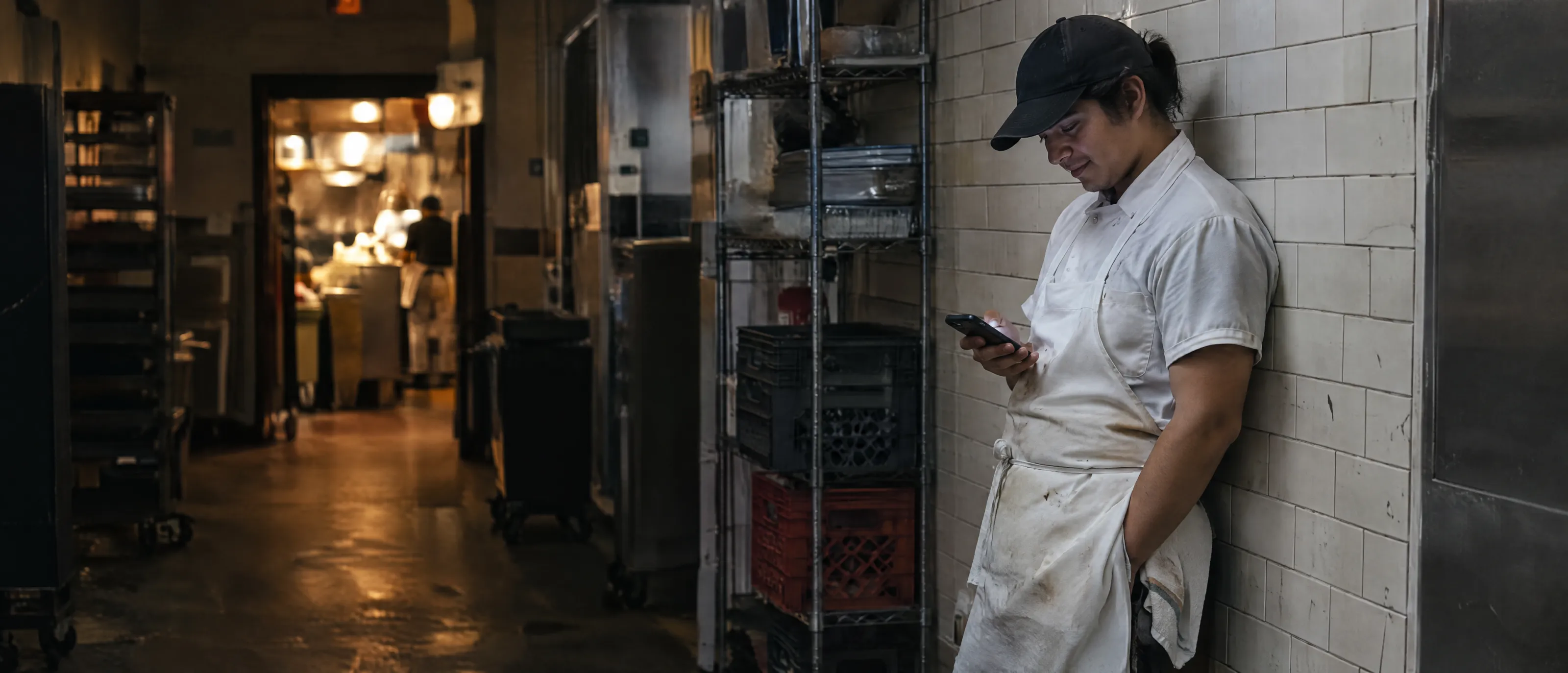 A line cook leaning on a tiled wall after the dinner rush, reading a text
